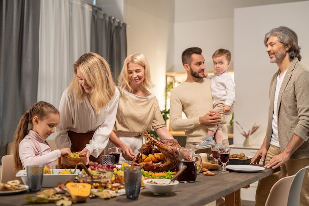 Young blond woman giving her little daughter fresh fruits while both standing by served table on background of the rest of family