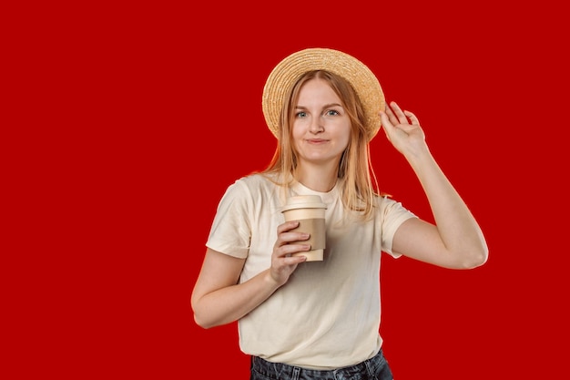 写真 赤い色の背景のスタジオ ポートレートに行く紙エコ コーヒーと帽子の若いブロンドの女性