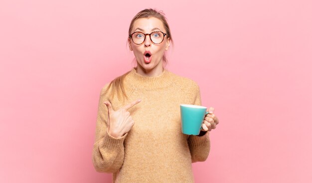 Young blond woman looking shocked and surprised with mouth wide open, pointing to self. coffee concept