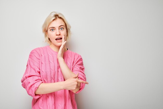 Young blond woman in pink sweater pointing aside finger demonstrating empty space for product or text isolated on white studio background