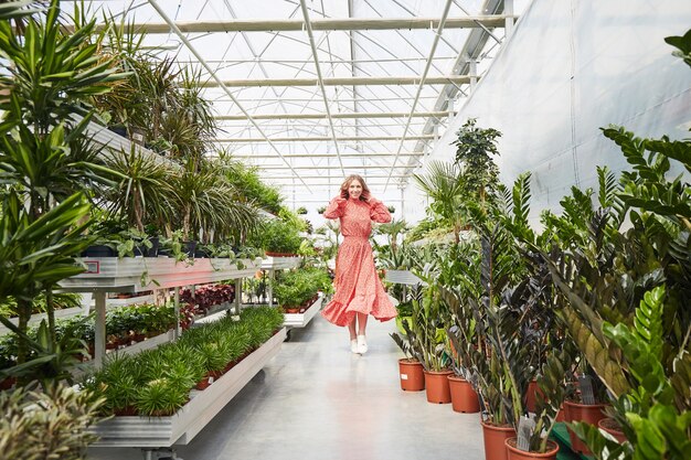 Young blond woman in red dress dancing into a greenhouse. Industrial Greenhouse with Flowers in pots all around.