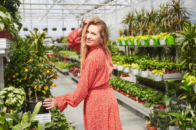 温室に赤いドレスを着た若いブロンドの女性。周りに鉢植えの花が咲く産業用温室。