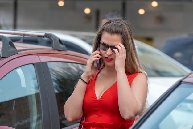 彼女の車の近くに赤いドレスを着て、電話で話している若いブロンドの女性現代のビジネスウーマン