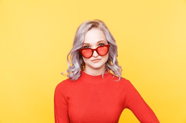 Young blond woman in red glasses and bright clothes watching camera.