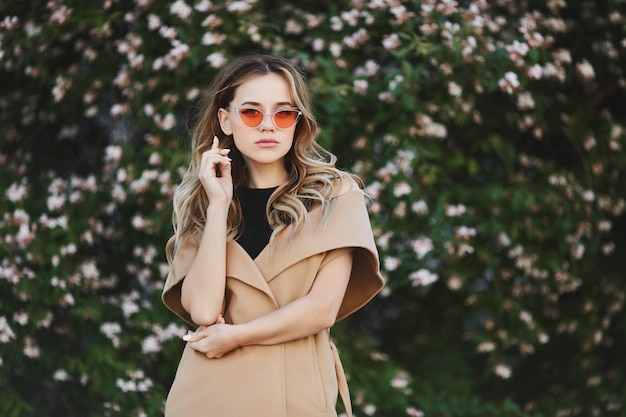 Young blond woman in sleeveless coat and in stylish sunglasses posing outdoors near blooming tree.