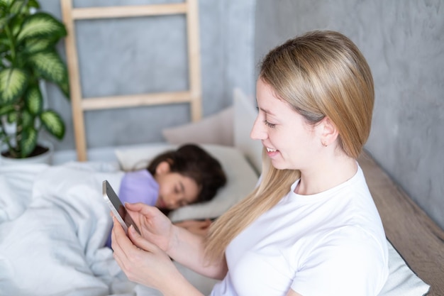 young blond woman using phone lying on bed with her sleeping cute child daughter