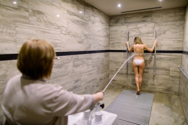 Young blond woman in white swimsuit having high pressure massage with Charcot shower for anti-cellulite treatment in the spa salon