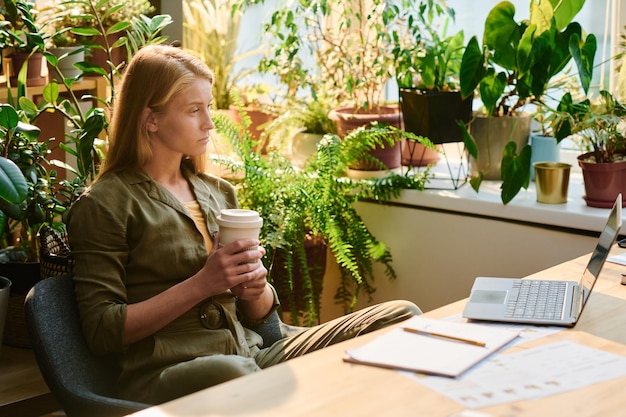 Young blond woman with cup of coffee looking at laptop screen\
during network