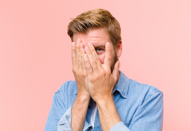 Young blonde adult man covering face with hands, peeking between fingers with surprised expression and looking to the side