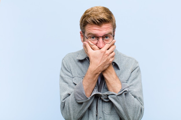 Young blonde adult man covering mouth with hands with a shocked, surprised expression, keeping a secret or saying oops