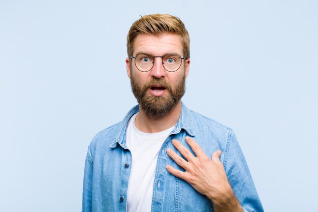 Young blonde adult man feeling shocked, astonished and surprised, with hand on chest and open mouth, saying who, me?