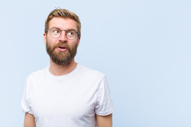 Young blonde adult man feeling shocked, happy, amazed and surprised, looking to the side with open mouth