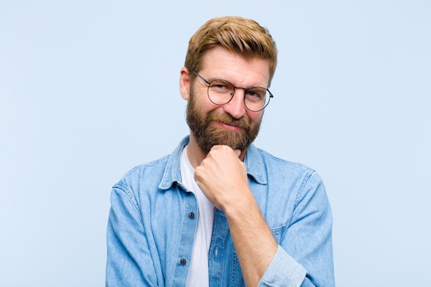 Young blonde adult man smiling, enjoying life, feeling happy, friendly, satisfied and carefree with hand on chin