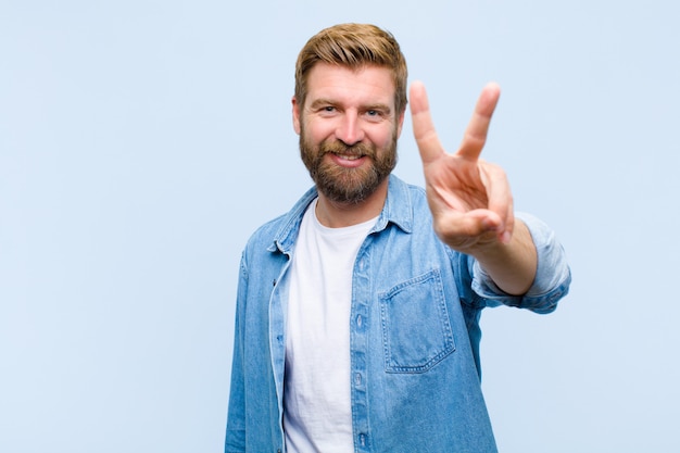 Young blonde adult man smiling and looking friendly, showing number two or second with hand forward, counting down