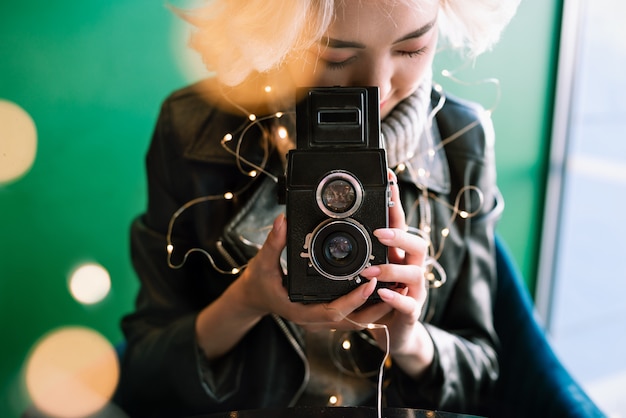Young blonde asian female holding vintage film camera with fairy lights and bokeh around, selective focus