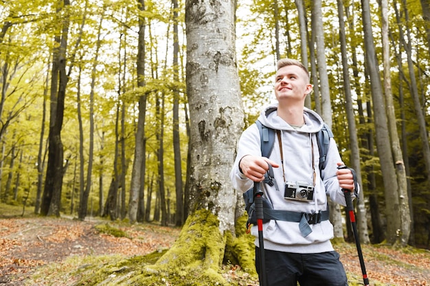 Young blonde attractive smiling man with trekking poles and camera standing in the forest and looking on the amazing nature views trekking recreation and healthy lifestyle concept