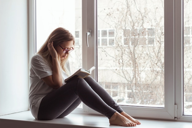 A young blonde beautiful woman in the white t-shirt and grey leggings with glasses is sitting on the windowsill and reading a book.