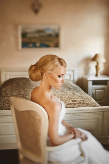 Young blonde beautiful woman with a perfect body and with stylish wedding hairstyle in a white dress sits in an armchair at the bedroom