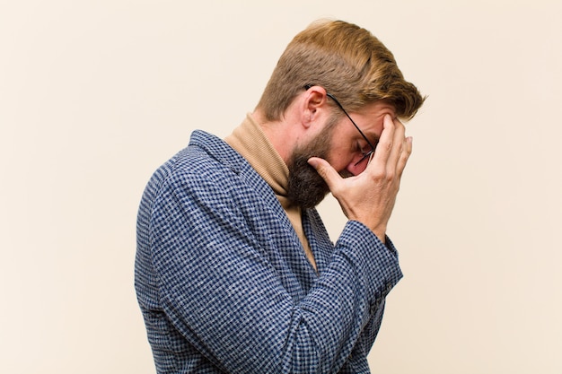 Young blonde businessman covering eyes with hands with a sad, frustrated look of despair, crying, side view