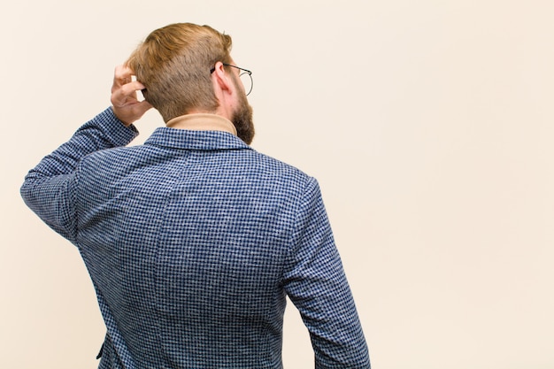 Young blonde businessman feeling clueless and confused, thinking a solution, with hand on hip and other on head, rear view