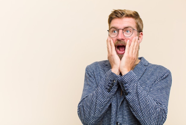 Young blonde businessman feeling happy, excited and surprised, looking to the side with both hands on face