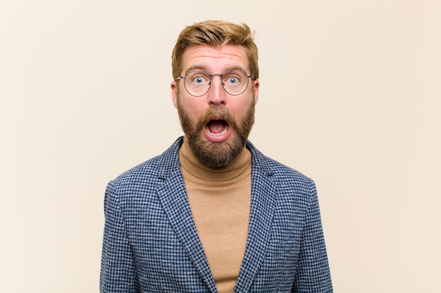 Young blonde businessman feeling terrified and shocked, with mouth wide open in surprise