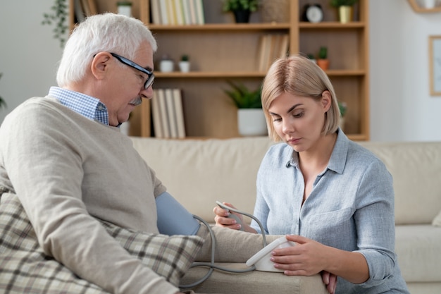 Young blonde careful woman with tonometer measuring blood pressure of her aged sick father at home