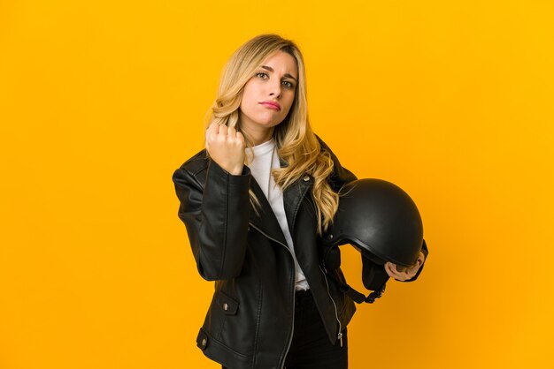 Young blonde caucasian biker woman holding helmet showing fist, aggressive facial expression.
