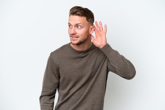 Foto giovane uomo biondo caucasico isolato su uno sfondo bianco che ascolta qualcosa mettendo la mano sull'orecchio