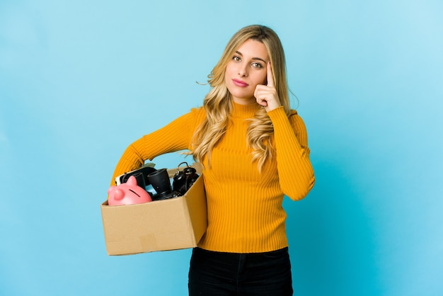 Young blonde caucasian woman holding boxes to move pointing temple with finger, thinking, focused on a task.