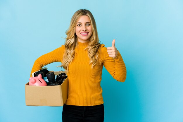 Young blonde caucasian woman holding boxes to move smiling and raising thumb up