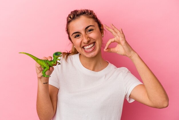 Giovane donna caucasica bionda che tiene una miniatura del dinosauro isolata su fondo rosa allegro e fiducioso che mostra gesto giusto.