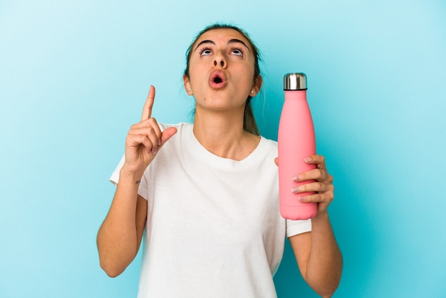 Young blonde caucasian woman holding a thermo isolated on blue wall pointing upside with opened mouth