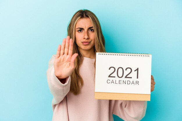 Young blonde caucasian woman holing a calendar isolated standing with outstretched hand showing stop sign, preventing you.