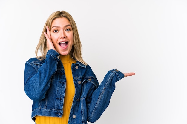 Young blonde caucasian woman isolated holds copy space on palm, keep hand over cheek. Amazed and delighted.