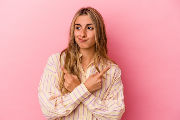 Young blonde caucasian woman isolated on pink wall points sideways, is trying to choose between two options