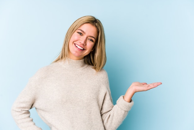 Young blonde caucasian woman isolated showing a copy space on a palm and holding another hand on waist.