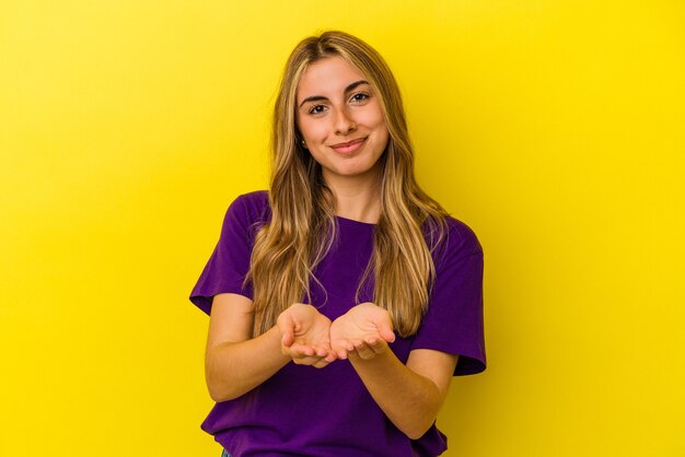 Giovane donna caucasica bionda isolata su fondo giallo che tiene qualcosa con le palme, offrendo alla macchina fotografica.