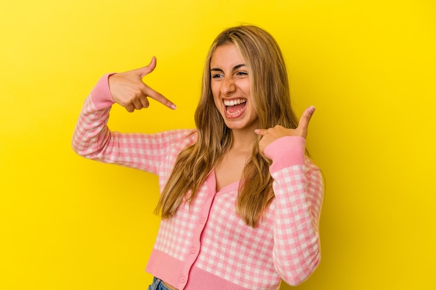 Young blonde caucasian woman isolated on yellow background surprised pointing with finger, smiling broadly.