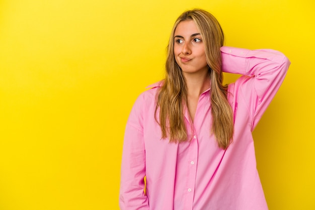 Young blonde caucasian woman isolated on yellow background touching back of head, thinking and making a choice