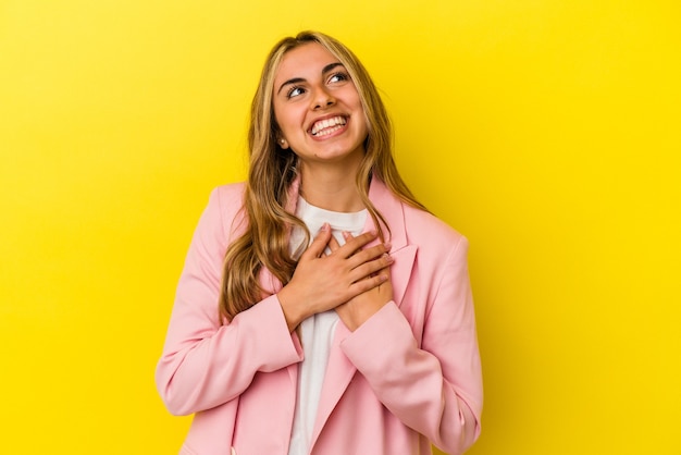 Young blonde caucasian woman isolated on yellow wall laughing keeping hands on heart, concept of happiness