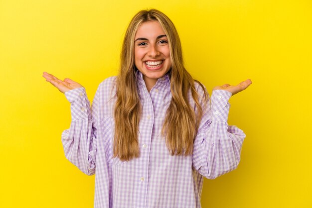 Young blonde caucasian woman isolated on yellow wall makes scale with arms, feels happy and confident