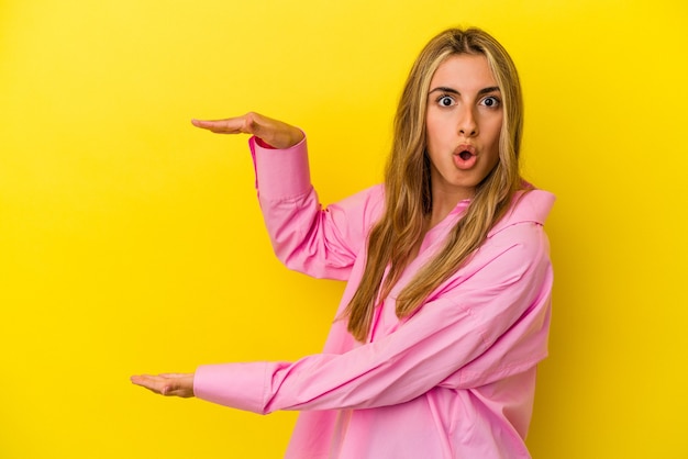 Young blonde caucasian woman isolated on yellow wall shocked and amazed holding a copy space between hands.