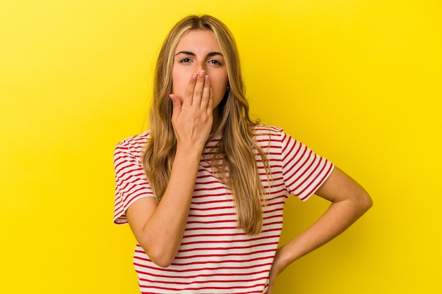 Giovane donna bionda caucasica isolata sulla parete gialla scioccata, che copre la bocca con le mani, ansiosa di scoprire qualcosa di nuovo.