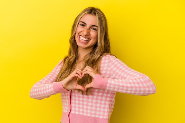 Young blonde caucasian woman isolated on yellow wall smiling and showing a heart shape with hands.