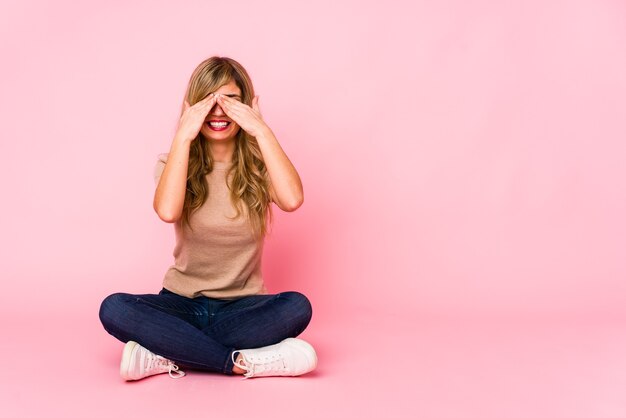 Young blonde caucasian woman sitting on a pink studio covers eyes with hands, smiles broadly waiting for a surprise.