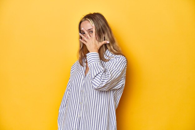 Young blonde Caucasian woman in a striped business shirt on a yellow background blink