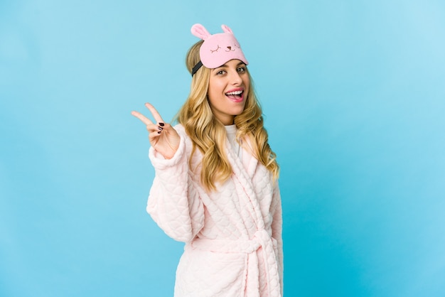 Young blonde caucasian woman wearing a pajama joyful and carefree showing a peace symbol with fingers.