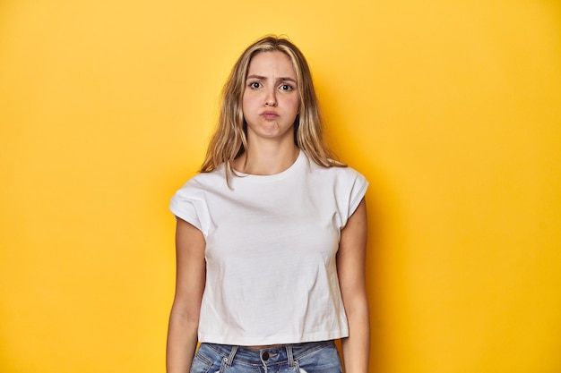 Foto giovane donna bionda caucasica con una maglietta bianca su uno sfondo giallo dello studio soffia le guance ha un'espressione stanca concetto di espressione facciale