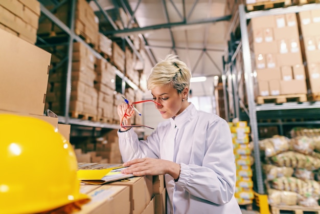 Young blonde Caucasian woman in white uniform and with eyeglasses filling out paperwork while standing in warehouse.